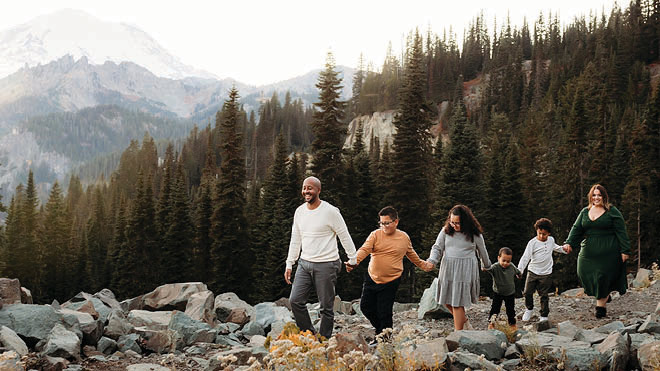 A family posing together in an outdoor setting, with mountain views, trees and nature all around them. The family of six is holding hands and standing in a line. 