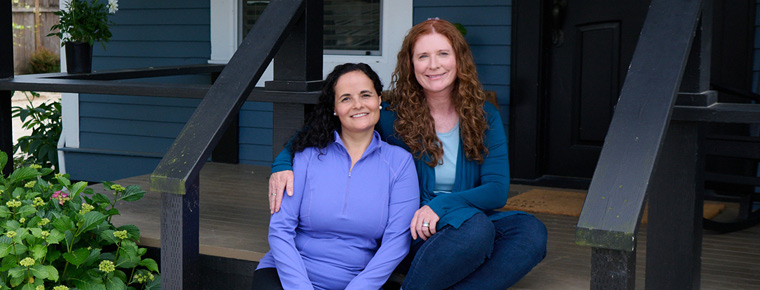 Two women sitting on the steps of a house. The woman on the right has one arm wrapped around the other. She has red long curly hair and is wearing a blue long sleeve cardigan and jeans. The woman on the left has black curly hair. She is wearing a periwinkle-colored long sleeve and black shorts. The home is blue with white trim around the windows and the steps are wooden. On the left are bushes with yellow flowers.