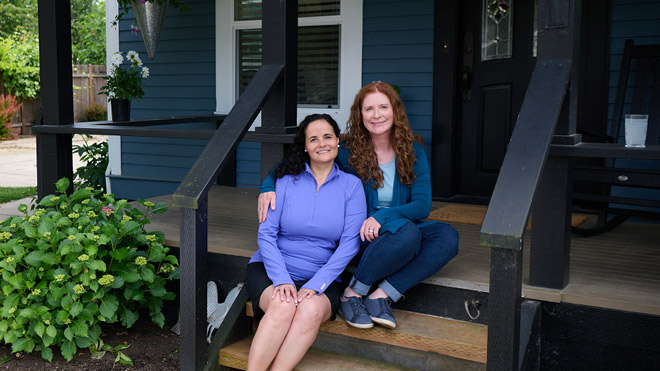 Two women sitting on the steps of a house. The woman on the right has one arm wrapped around the other. She has red long curly hair and is wearing a blue long sleeve cardigan and jeans. The woman on the left has black curly hair. She is wearing a periwinkle-colored long sleeve and black shorts. The home is blue with white trim around the windows and the steps are wooden. On the left are bushes with yellow flowers. 
