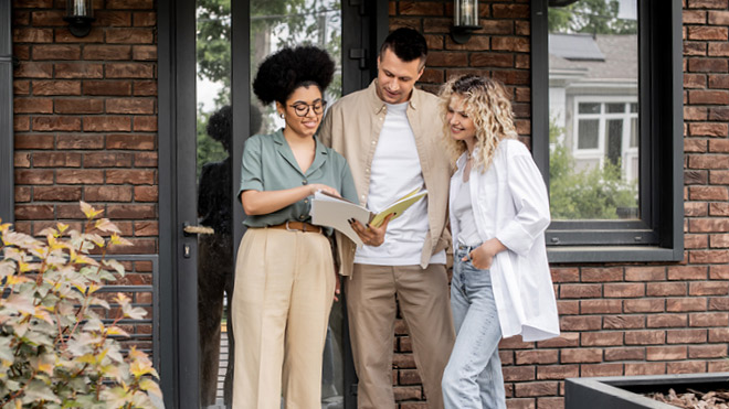 Three people standing in front of a brick house, one person is holding and showing a document to the other two. The woman holding the document has curly black hair and glasses. She's wearing a green top and beige pants. The couple, are standing next to each other, looking at the document. A man, in the middle, is wearing a beige top and brown pants. He has dark hair. The woman, to the right, is wearing a white top and light jeans. She has blonde hair. To the left are some plants.