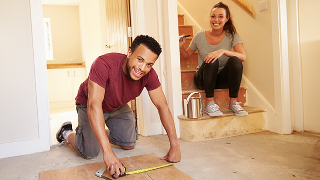 A man and woman engage in a flooring project. The man is kneeled over and is measuring flooring on the ground and the woman is sitting behind him on steps. There's a paint can on the steps next to the woman, She has a paint brush in her hand. Both of them are smiling.