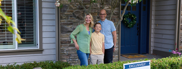 A family poses together in front of their house. Grouped to the left is a woman, in the middle is a child and to the right is a man. They are standing in the outside entryway in front of a green hedge and sold sign. 