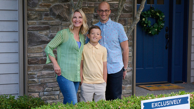 A family poses together in front of their house. Grouped to the left is a woman, in the middle is a child and to the right is a man. They are standing in the outside entryway in front of a green hedge and sold sign. 