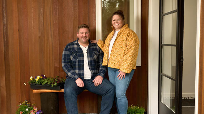 A man and woman pose together in front of the entryway of their home. The man is seated on a bench. On the bench are some potted flowers. The woman is standing next to him with her arm on his shoulder. They are in front of a rustic wooden background with a window behind them. To the right is the front door.