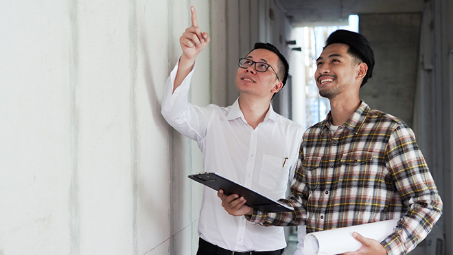Two men standing side by side, smiling as they collaborate on a home renovation project. The man on the left is pointing upward and wearing a white shirt. The main on the right is wearing a plaid shirt and is holding a clip board and rolled up sketch paper. They are in a hallway indoors.