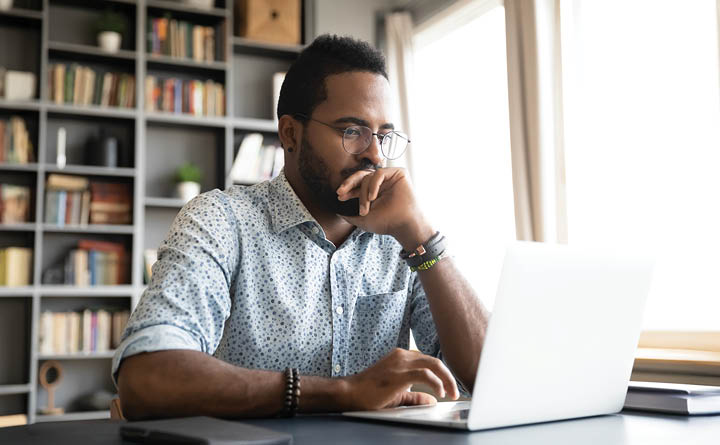 A man is seated at a desk, in a home office setting, focused on his laptop while working. He is wearing glasses and a button up shirt. In the background is a bookshelf with books and other home decor. The window next to him makes the setting bright. 