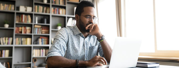 A man is seated at a desk, in a home office setting, focused on his laptop while working. He is wearing glasses and a button up shirt. In the background is a bookshelf with books and other home decor. The window next to him makes the setting bright. 