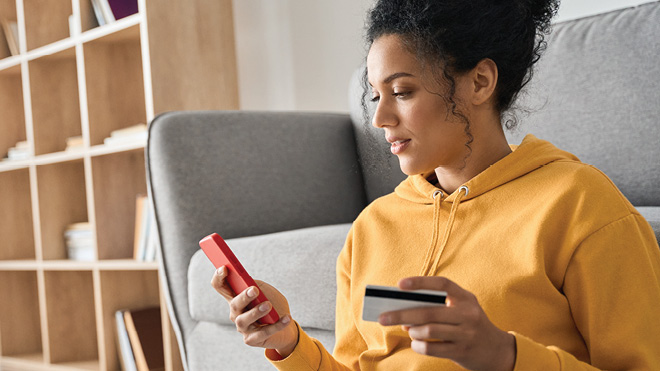 A woman in a yellow hoodie sits on the floor in a living room setting, holding a credit card and a smartphone, likely making an online purchase. There is a wooden bookshelf and gray couch in the background.