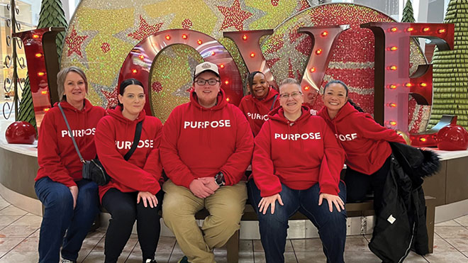 A group of BECU employees pose gather in front of a love sign in a festive mall setting. They are all wearing red sweatshirts that say "purpose."