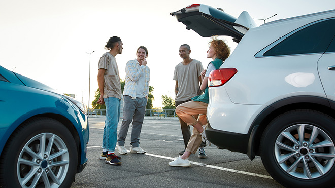 Four people gathered around an open vehicle trunk. They are chatting in a parking lot setting, having a conversation together.