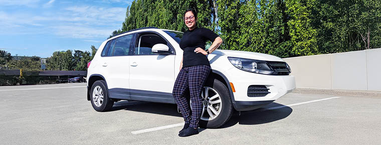 A woman stands next to a white SUV in a parking lot setting, she's wearing a black top and black plaid pants. She has one hand on her hip and is smiling proudly. There are trees and another parked car in the background.