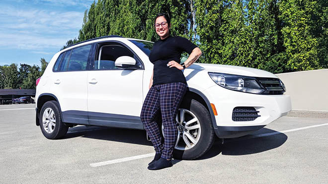  A woman stands next to a white SUV in a parking lot setting, she's wearing a black top and black plaid pants. She has one hand on her hip and is smiling proudly. There are trees and another parked car in the background.