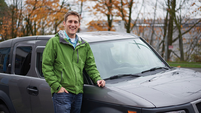  A man in a green jacket stands next to a gray SUV, he is wearing jeans and smiling. In the background are trees. 