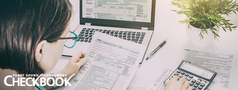An image of a person on a computer while filing their taxes. They are looking at what appears to be a W-2 Form. The form is in their left hand. The person is using their right hand to calculate numbers on a calculator. The Consumers' Checkbook logo is in the bottom left-hand side of the image. There is a pen on a white surfaced desk top and a plant. In the image you can see the back of the person's head and she is wearing blue glasses. 