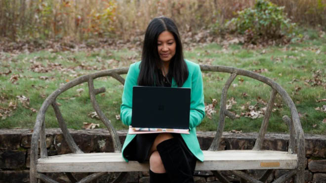 A woman on a bench, using a laptop, with a park setting in the background. She's wearing a teal blazer and has long dark hair.