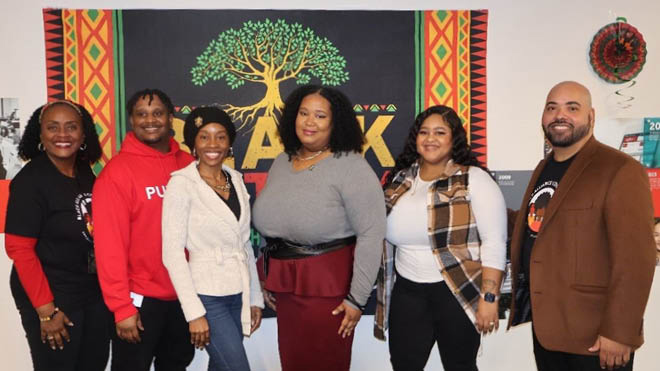 A group of BECU Black Alliance Cooperative (BAC) employee's pose in front of a Black History Month flag. The six of them are standing in a line and the flag consists of red, green, yellow and black colors with a tree in the middle.