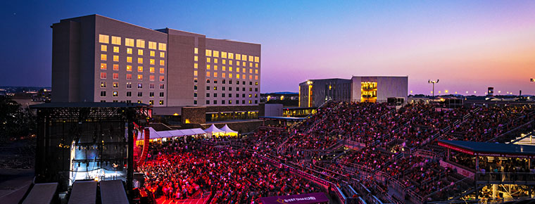 A large crowd gathers next to Northern Quest Casino for an outdoor concert. People are standing in bleachers and on the floor. Bright lights shine from the stage, and a sunset is visible in the background.