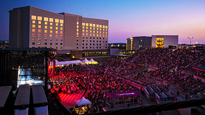 A large crowd gathers next to Northern Quest Casino for an outdoor concert. People are standing in bleachers and on the floor. Bright lights shine from the stage, and a sunset is visible in the background.