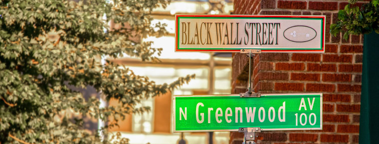 A green and white street sign. The green street sign says "Greenwood Ave" and the white sign says "Black Wall Street" with a key near the name. In the background is a brick building and trees. 