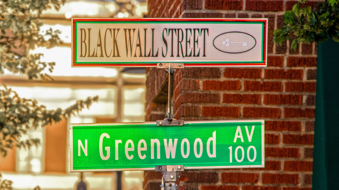A green and white street sign. The green street sign says "Greenwood Ave" and the white sign says "Black Wall Street" with a key near the name. In the background is a brick building and trees. 