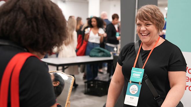 A smiling woman stands at a table with a promotional sign behind her. She has a light blue name badge and is wearing a black shirt. 