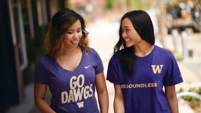 Two women walk side by side on a sidewalk, engaging in conversation. Both are wearing purple University of Washington t-shirts.