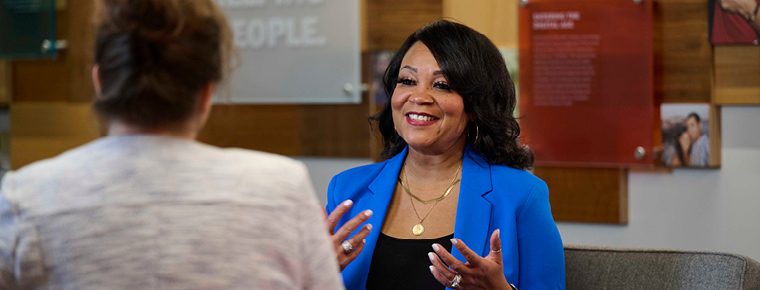 A woman in a blue blazer seated in a chair engages in conversation with another woman seated in front of her. The woman in blue has her hands up, like she's explaining a concept. They are in an indoor BECU financial setting.