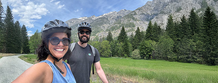  A couple in helmets biking on a trail, enjoying the outdoors together. In the background is a mountain view, trees and blue sky. The two are both wearing sunglasses.