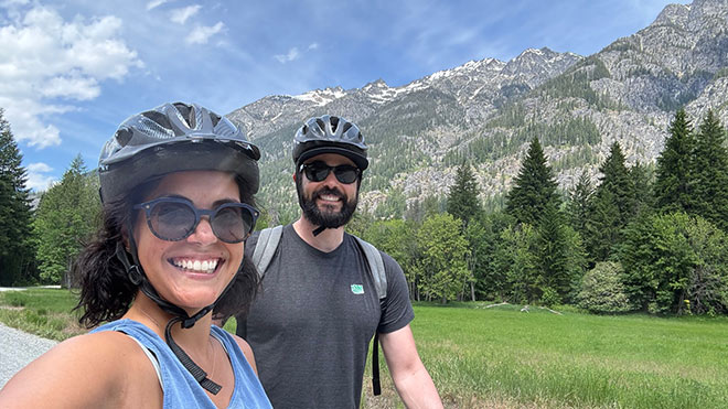  A couple in helmets biking on a trail, enjoying the outdoors together. In the background is a mountain view, trees and blue sky. The two are both wearing sunglasses.