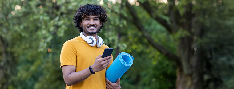 A man stands holding a blue yoga mat in one hand while smiling and holding his phone in the other hand. He's wearing a yellow t-shirt and is outdoors.