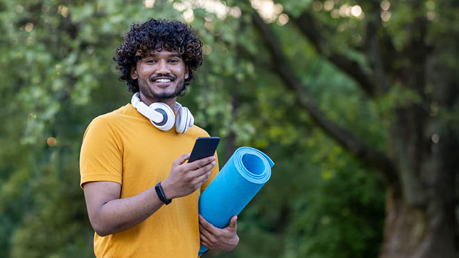 A man stands holding a blue yoga mat in one hand while smiling and holding his phone in the other hand. He's wearing a yellow t-shirt and is outdoors.