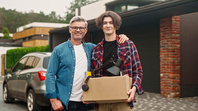 A father and son stand in front of a house. The son holds a box filled with personal belongings, suggesting he may be moving out.