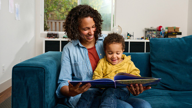 A mom and a child sitting on a couch, reading a book together. The two are smiling and both looking down at the book.