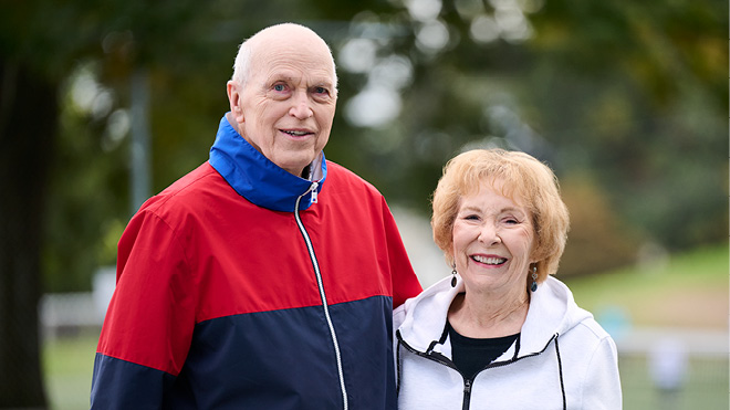 A senior couple stands closely in a park. There are trees and greenery in the background.