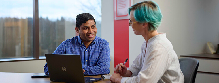 Two individuals seated at a table in an office setting, collaborating over a laptop. The woman, on the right, is holding a pen and has short blue hair. She's wearing a white top. The man, on the left, has dark hair and is wearing a blue long sleeve shirt.