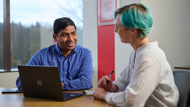 Two individuals seated at a table in an office setting, collaborating over a laptop. The woman, on the right, is holding a pen and has short blue hair. She's wearing a white top. The man, on the left, has dark hair and is wearing a blue long sleeve shirt.