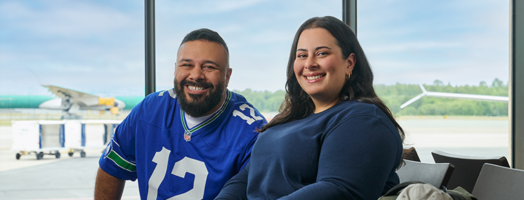 A couple seated in an airport terminal with a plane visible through the window behind them. The man wears a Seahawks 12th Man jersey, and the woman wears a blue sweater.