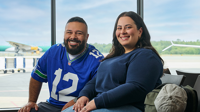 A couple seated in an airport terminal with a plane visible through the window behind them. The man wears a Seahawks 12th Man jersey, and the woman wears a blue sweater.