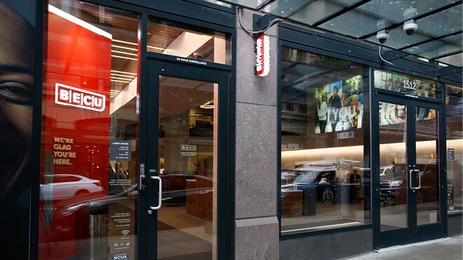 Exterior of a BECU neighborhood financial center with a red LED advertising screen visible. The interior of the building can be seen through the windows, and the building’s glass reflects cars passing on the street.