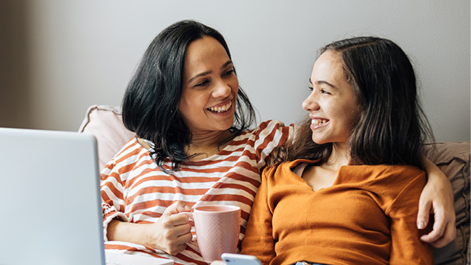 Mother and teenage daughter sitting on a couch, smiling and chatting while using a laptop.