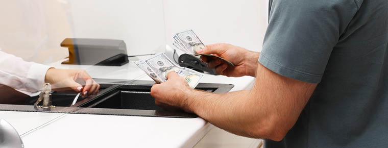 Close-up of a man's hands counting money while standing in a bank teller line. A woman's hands are visible behind the counter, as if she just gave him the money.