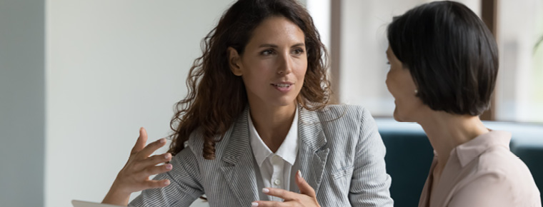 Two women in an office setting, one gesturing as if explaining something while the other listens attentively. A laptop sits on the desk in front of them.