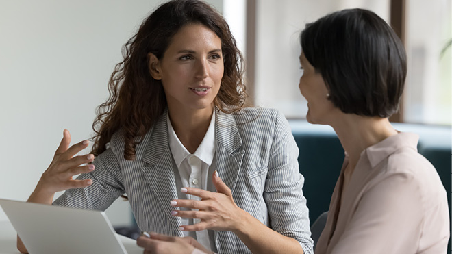 Two women in an office setting, one gesturing as if explaining something while the other listens attentively. A laptop sits on the desk in front of them.
