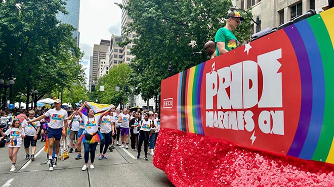 A big red-sided float carrying music DJs travels down a street in Seattle. The side has the BECU logo and says "Pride Marches On" in big white letters surrounded by rainbow colors. BECU employees are walking behind the float. Some are wearing rainbow clothes and carrying rainbow flags. 