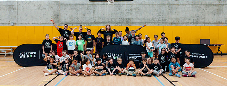 A diverse group of mostly children, along with a few adults in the back, pose and smile for a group photo in a gym. A basketball hoop and pop-up banners reading 'Together We Rise' and 'Presented by BECU and Starbucks' are visible in the background.