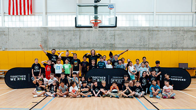 A diverse group of mostly children, along with a few adults in the back, pose and smile for a group photo in a gym. A basketball hoop and pop-up banners reading 'Together We Rise' and 'Presented by BECU and Starbucks' are visible in the background.