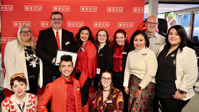 A group of BECU employees smiling in front of a red banner. There are about 11 employees in the photo.