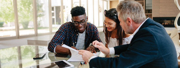 Three people sit at a kitchen island with documents and a laptop, discussing a possible sale. One man appears to be signing papers while his partner looks at the papers and smiles.