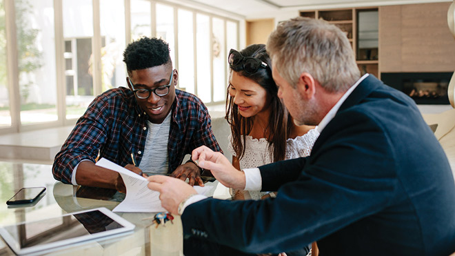 Three people sit at a kitchen island with documents and a laptop, discussing a possible sale. One man appears to be signing papers while his partner looks at the papers and smiles.