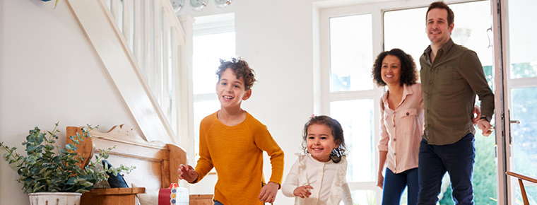 A family of four enters a home with excitement. The parents walk arm in arm, with the father holding the open door. Their two smiling children lead the way into the entryway.
